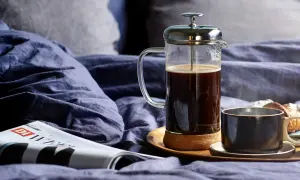 En franskpress med kaffe står på en bricka i en säng. Bredvid finns en kopp, en tidning och en croissant. Perfekt för en lugn morgon, men hur många matskedar kaffe per kopp är lagom?