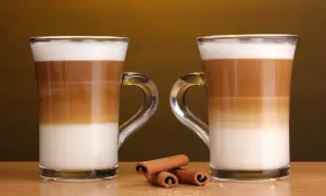 Två glas med lager av mjölk och kaffe, en perfekt jämförelse mellan latte macchiato vs caffe latte. Kanelstänger ligger framför.