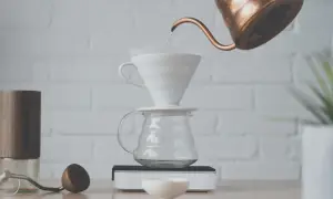 Vatten hälls från en kopparkanna över ett filter i en vit keramiktratt, som står på en glaskaraff. En kaffekvarn och en träsked ligger bredvid. Det är dags att brygg.