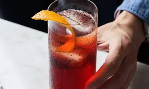 Ett glas med iskallt, rött americano kaffe, garnerat med en apelsinzest. En hand sträcker sig efter drinken.