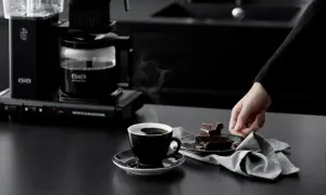 En hand sträcker sig efter chokladbitar bredvid en kopp ångande kaffe, nyligen bryggd med Moccamaster.