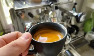 En hand håller en kopp med gyllene spanskt kaffe framför en espressomaskin.