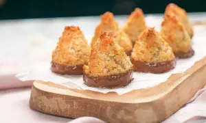 Små, gyllene kokoskakor glutenfria, doppade i choklad, ligger på ett vitt papper på en träbricka.
