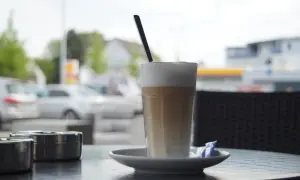 En hög latte macchiato med skummjölk och en sked på ett fat, bredvid två askkoppar.