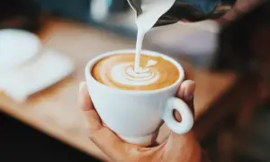 En hand håller en vit kopp med en vacker kaffe latte, där mjölkskummet skapar ett hjärta.