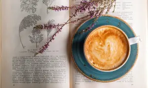 En kopp kaffe med lattekonst vilar på en öppen bok med botaniska illustrationer. En lila blomsterkvist ligger över boken och koppen, som om den vore en del av en studie för att inte frysa kaffe.