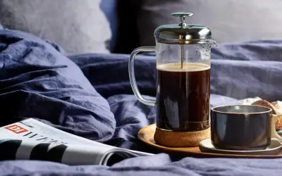 En franskpress med kaffe står på en bricka i en säng. Bredvid finns en kopp, en tidning och en croissant. Perfekt för en lugn morgon, men hur många matskedar kaffe per kopp är lagom?