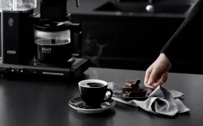 En hand sträcker sig efter chokladbitar bredvid en kopp ångande kaffe, nyligen bryggd med Moccamaster.