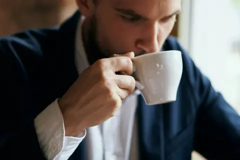 En man i kostym tar en klunk kaffe ur en vit kopp. Kanske blir man pigg av kaffe.
