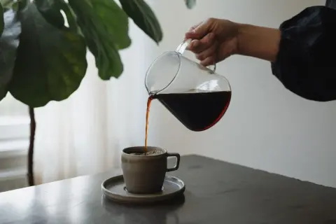 En hand häller upp mörkt kaffe från en glaskanna i en mugg. En fin stund för att njuta av kaffe, inte espresso.
