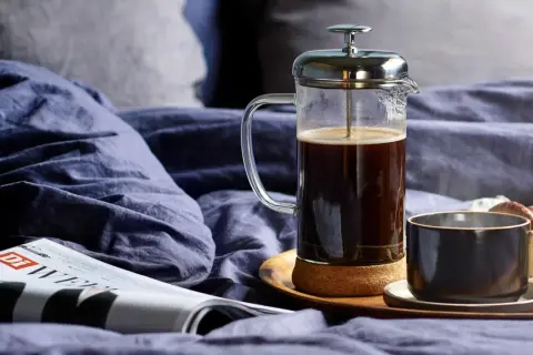 En franskpress med kaffe står på en bricka i en säng. Bredvid finns en kopp, en tidning och en croissant. Perfekt för en lugn morgon, men hur många matskedar kaffe per kopp är lagom?