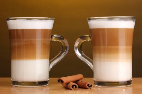 Två glas med lager av mjölk och kaffe, en perfekt jämförelse mellan latte macchiato vs caffe latte. Kanelstänger ligger framför.