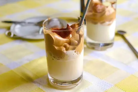 Två glas med krämigt dalgona iskaffe, toppat med chokladpulver, står på en gulrutig duk.
