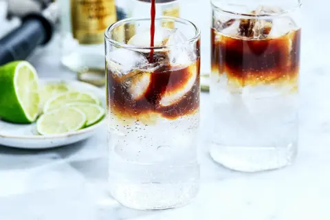 Två glas med kaffe tonic, is och lime. En flaska och en limeklyfta i bakgrunden.