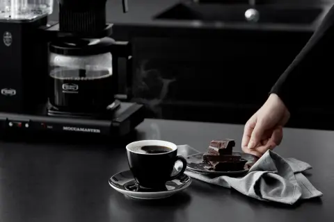 En hand sträcker sig efter chokladbitar bredvid en kopp ångande kaffe, nyligen bryggd med Moccamaster.