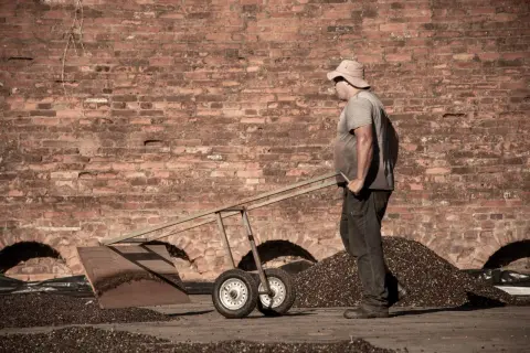 Man med hatt skottar kaffebönor framför en tegelvägg. Kanske en scen från vilket land producerar mest kaffe.