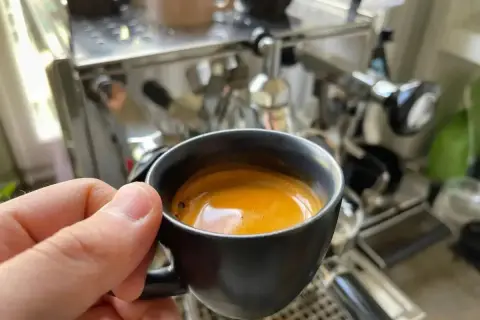En hand håller en kopp med gyllene spanskt kaffe framför en espressomaskin.