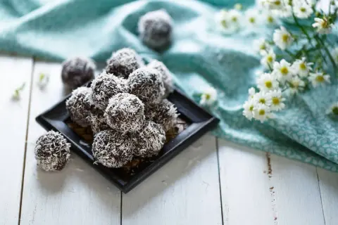 En hög med chokladbollar, perfekta för chokladbollens dag, rullade i kokosflingor. Blommor och en turkos duk i bakgrunden.