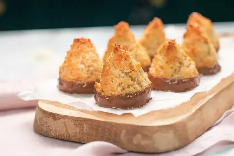 Små, gyllene kokoskakor glutenfria, doppade i choklad, ligger på ett vitt papper på en träbricka.