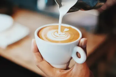 En hand håller en vit kopp med en vacker kaffe latte, där mjölkskummet skapar ett hjärta.
