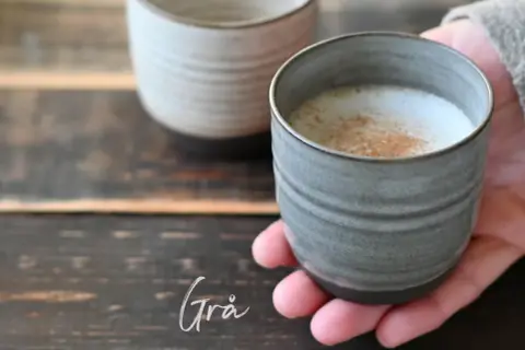En hand håller en grå mugg med ett skummande, kaneldoftande lungo kaffe. En annan mugg syns i bakgrunden.