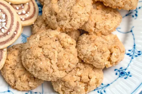 En hög med hembakade havrekakor med kokos, gyllene och krispiga, ligger på en tallrik med blått mönster.