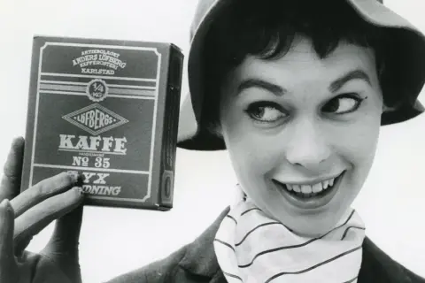 En kvinna med hatt och randig scarf håller upp en låda Löfbergs Kaffe. Kanske hon vet vilka som dricker mest kaffe i Sverige?
