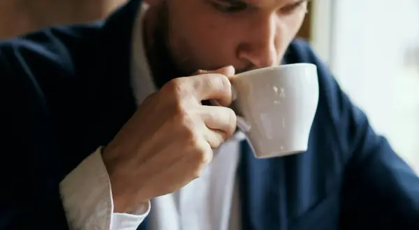 En man i kostym tar en klunk kaffe ur en vit kopp. Kanske blir man pigg av kaffe.