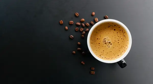 En kopp kaffe med skummig yta och kaffebönor runt omkring. Kanske någon som har en vegan i kaffet idag?