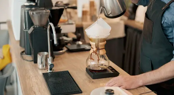 Barista häller vatten över kaffefilter i en Chemex. En kaffekvarn och en kran syns i bakgrunden. Intressant kaffe fakta!