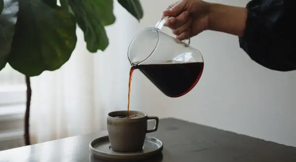 En hand häller upp mörkt kaffe från en glaskanna i en mugg. En fin stund för att njuta av kaffe, inte espresso.