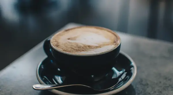 En kopp cappuccino latte med vacker skumkonst på en mörk tallrik med en sked.