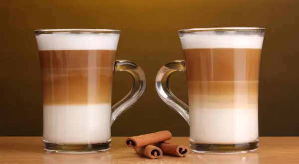 Två glas med lager av mjölk och kaffe, en perfekt jämförelse mellan latte macchiato vs caffe latte. Kanelstänger ligger framför.