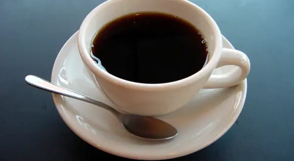 En vit kopp med mörkt kaffe och en sked på en fat. En påminnelse om kaffe ursprung och dess resa till din kopp.