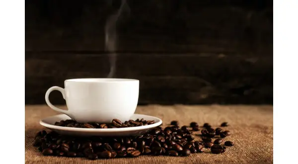 En vit kaffekopp med ånga stiger från den, omgiven av kaffebönor. En perfekt bild för att fundera på hur många skopor kaffe till 2 koppar.