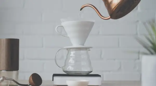 Vatten hälls från en kopparkanna över ett filter i en vit keramiktratt, som står på en glaskaraff. En kaffekvarn och en träsked ligger bredvid. Det är dags att brygg.