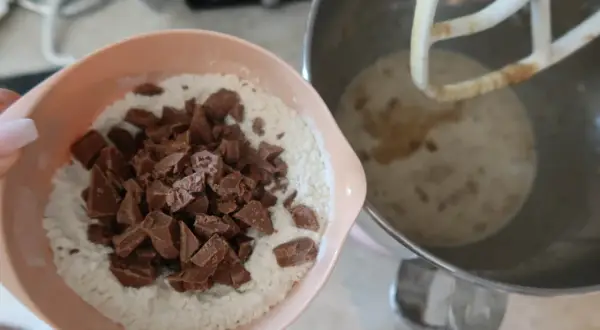 En skål med mjöl och chokladbitar för en saftig banankaka med choklad. Bredvid står en köksmaskin med smet.