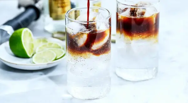 Två glas med kaffe tonic, is och lime. En flaska och en limeklyfta i bakgrunden.