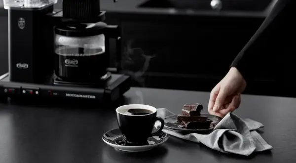 En hand sträcker sig efter chokladbitar bredvid en kopp ångande kaffe, nyligen bryggd med Moccamaster.