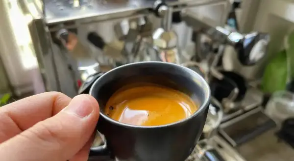 En hand håller en kopp med gyllene spanskt kaffe framför en espressomaskin.