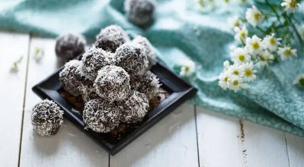 En hög med chokladbollar, perfekta för chokladbollens dag, rullade i kokosflingor. Blommor och en turkos duk i bakgrunden.