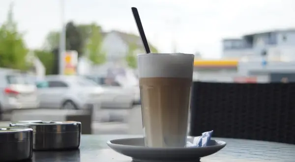 En hög latte macchiato med skummjölk och en sked på ett fat, bredvid två askkoppar.