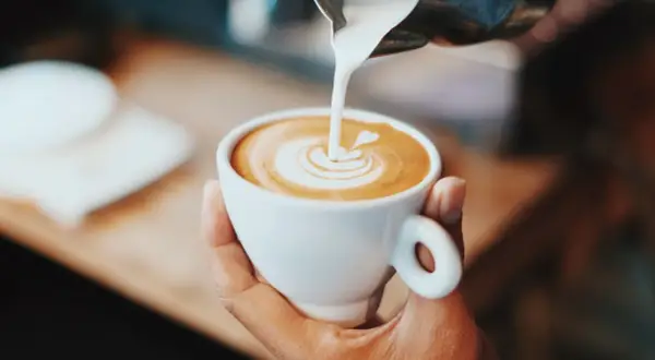 En hand håller en vit kopp med en vacker kaffe latte, där mjölkskummet skapar ett hjärta.