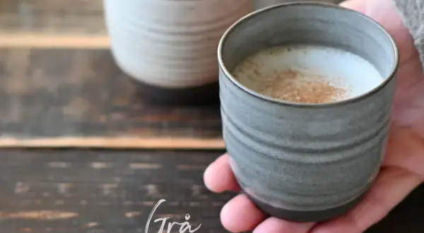 En hand håller en grå mugg med ett skummande, kaneldoftande lungo kaffe. En annan mugg syns i bakgrunden.