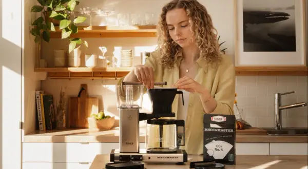 En kvinna förbereder starkt bryggkaffe med en Moccamaster. Solen lyser in i köket.