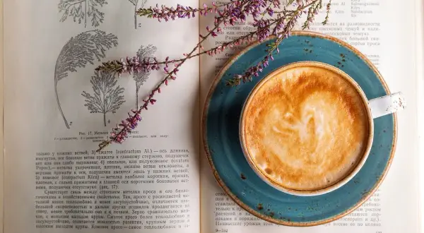 En kopp kaffe med lattekonst vilar på en öppen bok med botaniska illustrationer. En lila blomsterkvist ligger över boken och koppen, som om den vore en del av en studie för att inte frysa kaffe.