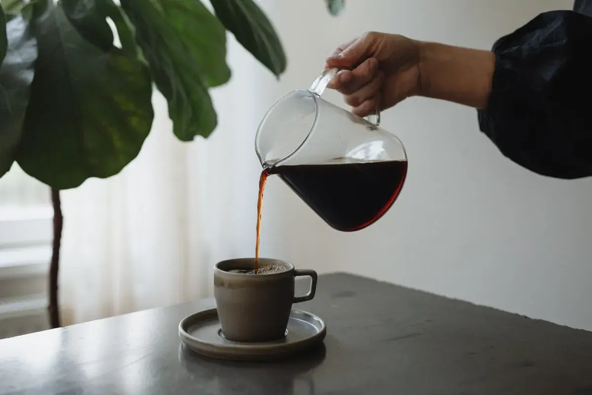En hand häller upp mörkt kaffe från en glaskanna i en mugg. En fin stund för att njuta av kaffe, inte espresso.