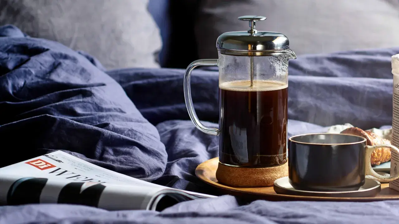 En franskpress med kaffe står på en bricka i en säng. Bredvid finns en kopp, en tidning och en croissant. Perfekt för en lugn morgon, men hur många matskedar kaffe per kopp är lagom?