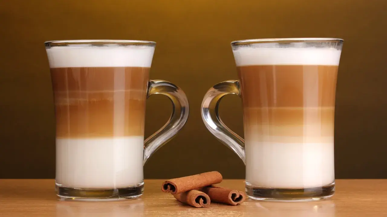 Två glas med lager av mjölk och kaffe, en perfekt jämförelse mellan latte macchiato vs caffe latte. Kanelstänger ligger framför.