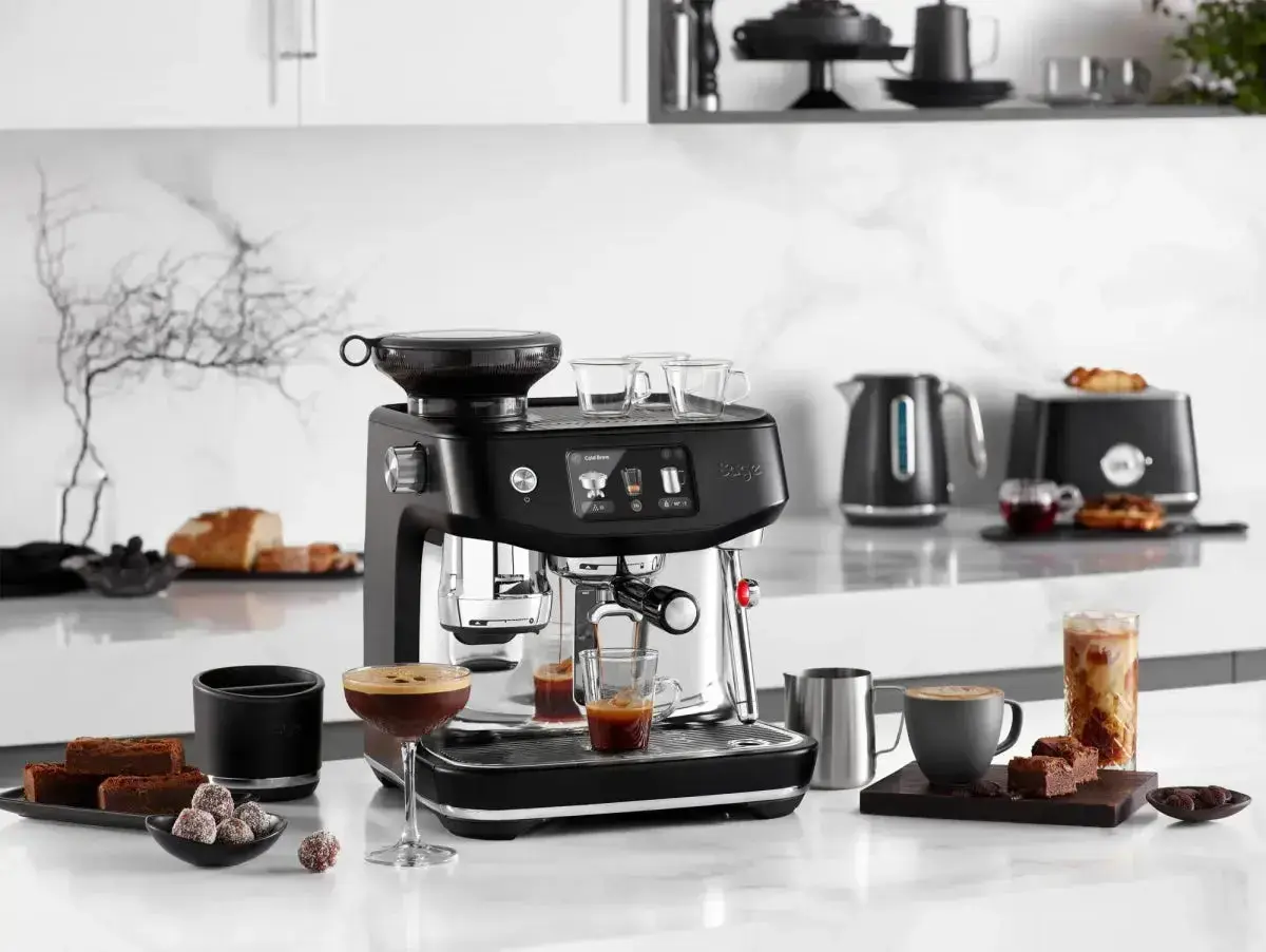 Svart espressomaskin med kaffekoppar, chokladbitar och en glasburk med iskaffe. Perfekt för att brygga gott kaffe oavsett om du har hårt eller mjukt vatten.